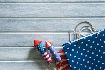 4th July holiday shopping concept. Shopping bags in national american colors and firework rockets over wooden background