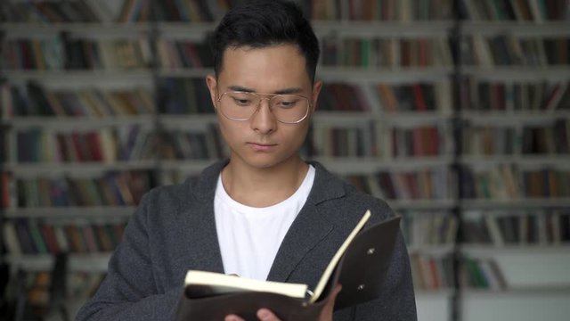 Close up portrait of black hair asian young man wearing grey jacket in eyeglasses standing alone reading flipping through a blocknote on background of bookshelves