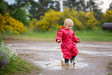 Fototapeta premium Sweet little toddler boy, blond child in red raincoat and blue boots, playing in the rain in muddy puddles, jumping and laughing