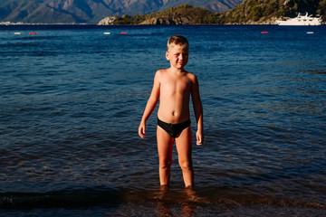 happy tanned seven-year-old boy in bathing trunks standing in the sea in Turkey and looking at the camera.