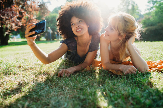 Two Young Women Takes A Selfie With The Smart Phone - Millennial Relaxing At The Park At Sunset On A Summer Day