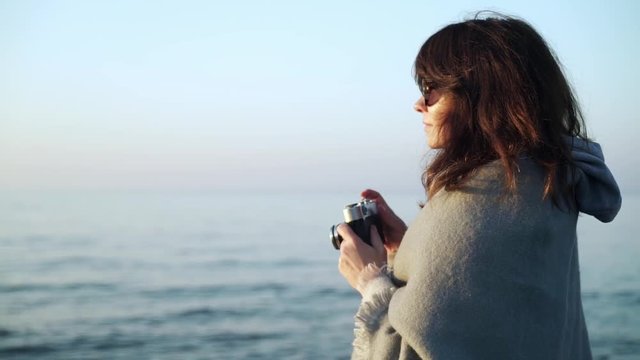 Slow motion of caucasian young woman wearing grey scarf and sunglasses taking photos with film camera of ocean waves on a cold day
