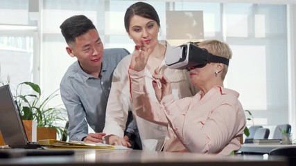 Mature businesswoman using 3d virtual reality goggles at work. Diverse business team discussing modern gadgets, female entrepreneur talking to colleagues, using vr glasses
