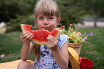 little beautiful girl of 4 years old in a dress and hat is eating a watermelon in the summer in the park at a picnic and smiling