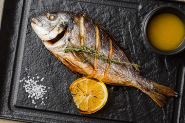 Delicious grilled dorado or sea bream fish with lemon slices, spices, rosemary on dark stone....