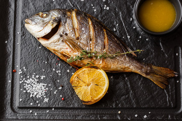 Delicious grilled dorado or sea bream fish with lemon slices, spices, rosemary on dark stone. Grilled sea fish with olive oil, spices and lemon ready for eating. Dorado, herbs and spices. Menu photo