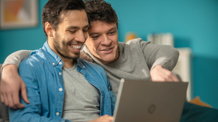 Adorable Male Gay Couple Spend Time at Home. They Sit on a Sofa and Use the Laptop. They Watch Funny Online Videos. Partner Puts His Hand Around His Lover. Room Has Modern Interior.