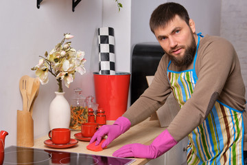 Young handsome bearded man in the kitchen, wearing apron and pink gloves cleans the cooking surface using detergents