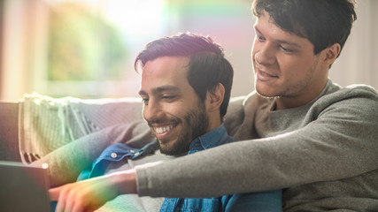 Male Queer Couple Spend Time at Home. They are Lying Down on Sofa and Use Laptop. They Browse Online. Partner's Hand is Around His Lover and Pointing on Screen. Shot with Rainbow Lens Flare Effect.