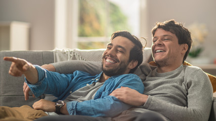 Sweet Male Queer Couple Spend Time at Home. They are Lying Down on a Sofa and Laughing. Partner's Hand is Around His Lover and Other Pointing on Screen. Room Has Modern Interior. - obrazy, fototapety, plakaty