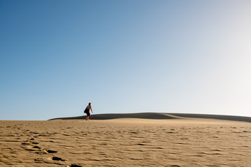 Fototapeta na wymiar evocative young confident woman walking on her own path on the desert sand with red dress in the middle of dunes on hot summer day with clear blue sky