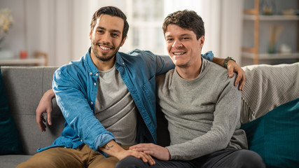 Cute Attractive Male Gay Couple Sit Together on a Sofa at Home. Boyfriend Puts His Hand on Partner's. They are Happy and Smiling. They are Casually Dressed and Their Room Has Modern Interior.