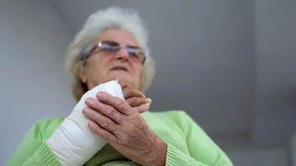 Sad old woman having pain sitting and holding her injured hand in bandages