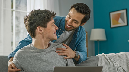Obraz premium Sweet Male Gay Couple Spend Time at Home. Young Man Works on a Laptop, His Partner Comes From Behind and Gently Embraces Him. They Laugh. Room Has Modern Interior.