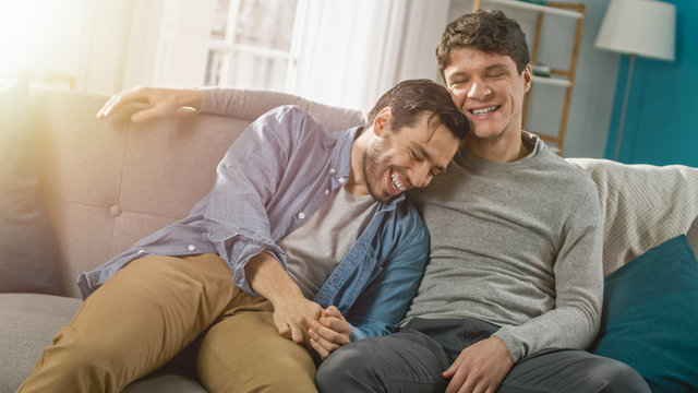Cute Attractive Male Gay Couple Sit Together on a Sofa at Home. Boyfriends are Hugging and Embracing Each Other. They are Joyful and Laughing. They are Casually Dressed and Room Has Modern Interior.