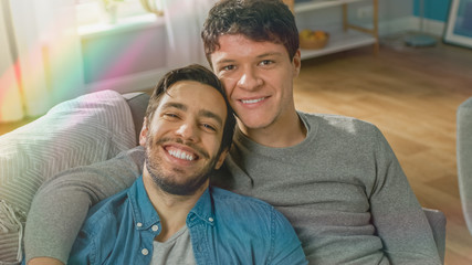 Portrait of Cute Male Queer Couple at Home. They Sit on Sofa and Look at Camera. Partner Embraces His Lover from Behind. They are Happy and Smiling. Room Has Modern Interior with Rainbow Flare Effect