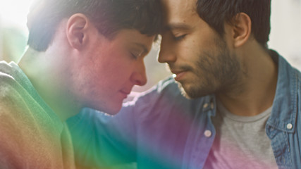 Cute Male Queer Couple Put Their Heads Together and Close Their Eyes. Sweet Gentle Loving Gay Relationship Moment. Room Has Modern Interior. Shot with Rainbow Flare Effect.