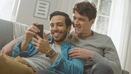 Fototapeta premium Cute Male Gay Couple Spend Time at Home. They are Lying Down on a Sofa and Use a Smartphone. They Browse Online. Partner's Hand is Around His Lover. They Smile and Laugh. Room Has Modern Interior.