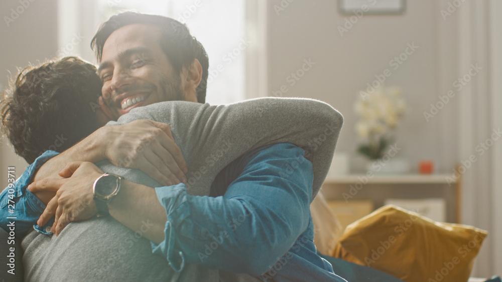 Wall mural cute attractive male gay couple sit together on a sofa at home. boyfriends are hugging and embracing