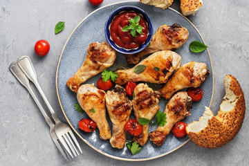 Closeup of fried chicken drumstick with tomatoes, ketchup and fresh bagel bread over gray background. Top view, flat lay.