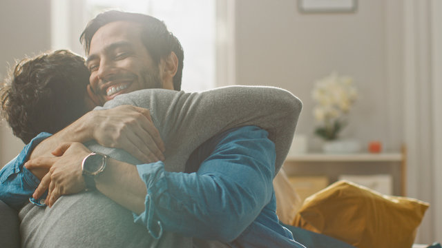 Cute Attractive Male Gay Couple Sit Together on a Sofa at Home. Boyfriends are Hugging and Embracing Each Other. They are Joyful and Laughing. They are Casually Dressed and Room Has Modern Interior. - Powered by Adobe