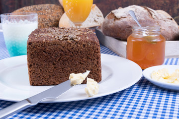 Multigrain bread with sunflower seeds served on a white plate, a jar of jam and loaves of bread in a basket.