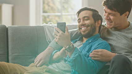 Cute Male Gay Couple Spend Time at Home. They are Lying Down on a Sofa and Use a Smartphone. They Browse Online. Partner's Hand is Around His Lover. They Smile and Laugh. Room Has Modern Interior.