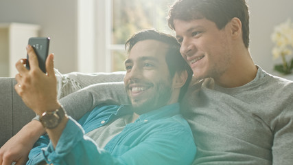 Cute Male Gay Couple Spend Time at Home. They are Lying Down on a Sofa and Use a Smartphone and Making a Selfie. They Browse Online. Partner's Hand is Around His Lover. They Smile and Laugh.