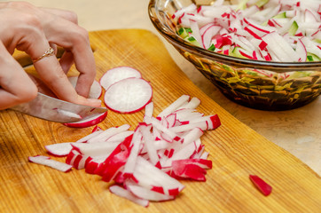 Female hands cut radish into small pieces on a wooden board close-up, next to it stands a glass brown salad bowl