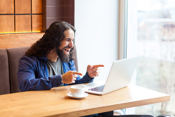 Portrait of happy suprised handsome young adult man freelancer in casual style sitting in cafe and watching funny video in laptop, toothy smile, pointing two finger to screen. Indoor,lifestyle concept