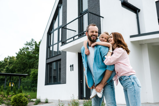 happy bearded man piggybacking daughter near cheerful wife and home