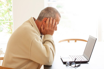 Casual dressed man sitting by a laptop computer is hiding his head in despair. A pair of glasses lying on a pile of papers next to the laptop. - Image