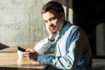 Side view portrait of satisfied attentive young bearded man in blue denim shirt sitting in cafe and holding phone and thinking. have a rest after work, looking at camera, indoor, lifestyle