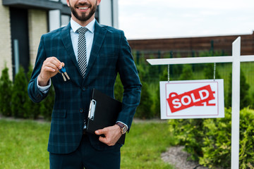 cropped view of cheerful broker holding keys and clipboard near board with sold letters
