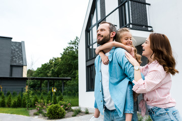 happy man piggybacking daughter near cheerful wife and house