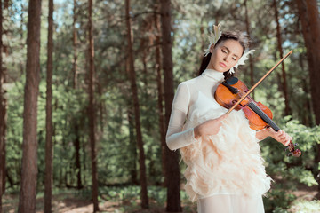 dreamy woman in white swan costume standing on forest background, playing on violin