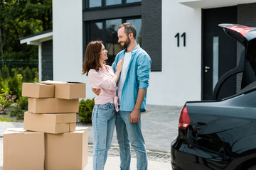 happy woman standing with bearded man near boxes and new house - obrazy, fototapety, plakaty