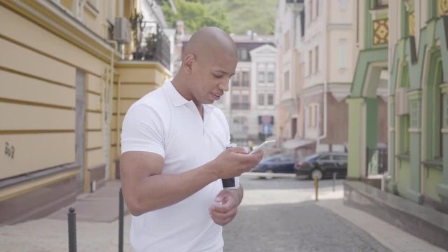 Portrait of handsome successful confident bald middle eastern man typing on the cell phone standing on the street in front of old buildings looking around. Professional skills.