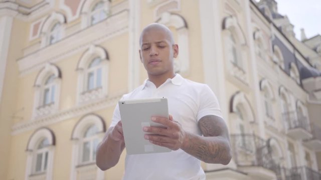 Portrait handsome bald middle eastern businessman typing on the tablet tanding on the city street. Freelance concept. Man working outdoors.