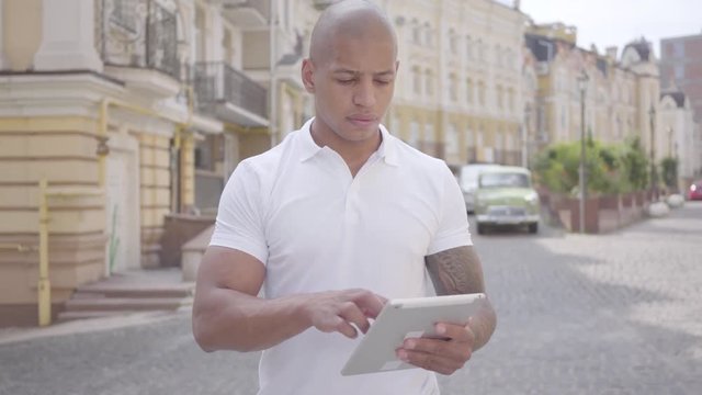 Handsome bald middle eastern businessman typing on the tablet tanding on the city street. Freelance concept. Man working outdoors.