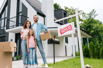 happy man holding box and standing with wife and daughter near house and board with sold letters