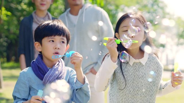 two little asian children boy and girl playing outdoors blowing soap bubbles with parents watching from behind.