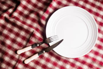 Empty white plate, fork and knife on a checkered red napkin, top view. Image with copy space. Kitchen table with a towel and a plate - top view with copy space. 