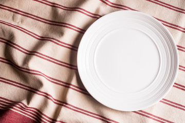 Empty white plate on a red striped napkin on an old wooden brown background, top view. Image with copy space. Kitchen table with a towel and a plate - top view with copy space. 