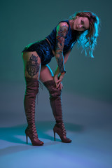 Attractive young blonde girl in blue velour booty shorts and maroon suede high boots posing on camera in studio