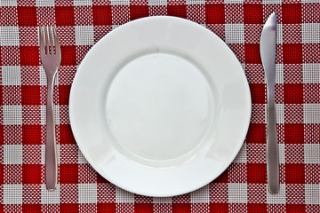white plate without food with a fork and knife on a wicker red and white tablecloth