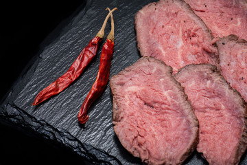 Sliced Grass Fed Juicy Corn Roast Beef garnished with dried Red Chile De Arbol Pepper on black natural stone background.