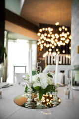 Wedding table setting is decorated with fresh flowers and white candles. Wedding floristry. Bouquet with roses, hydrangea and eustoma. On the background blur are burning garlands with light bulbs.