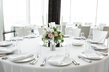Wedding table setting decorated with fresh flowers. White plates, silverware, white tablecloth and white room. Wedding floristry. Banquet table for guests. Bouquet with roses, hydrangea and eustoma