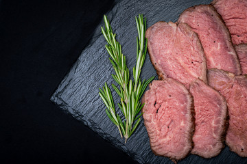Sliced Grass Fed juicy Corn Roast Beef garnished with Rosemary Fresh Herb on natural black stone background.